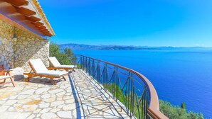 A stone balcony with lounge chairs overlooking a clear blue sea and distant mountains. - House On The Rocks in Nisaki (Corfu)