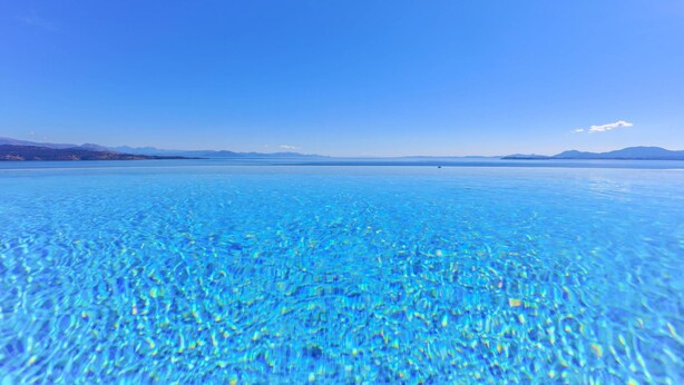 A clear view of a large, calm body of water with a distant mountain range under a blue sky. - House On The Rocks in Nisaki (Corfu)