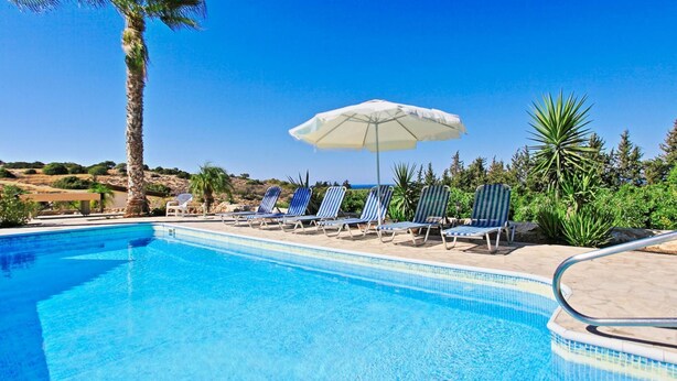 Ein Poolbereich mit Liegestühlen und Sonnenschirm, umgeben von Grünpflanzen und blauem Himmel.