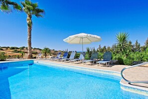 A pool area with lounge chairs and an umbrella, surrounded by greenery and a clear sky.