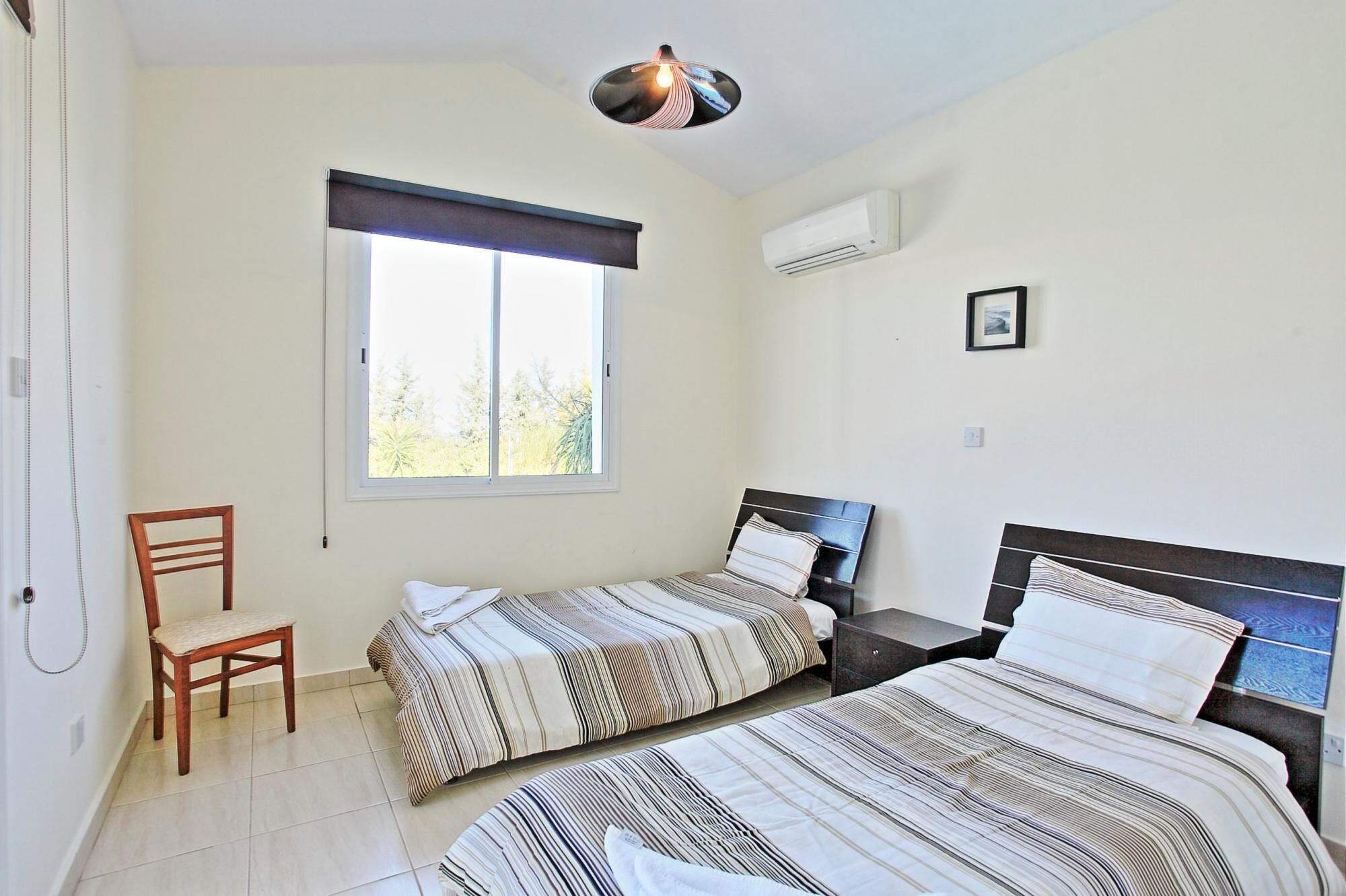 Une chambre avec deux lits, une chaise, une fenêtre, un appareil de climatisation et un tableau au mur.