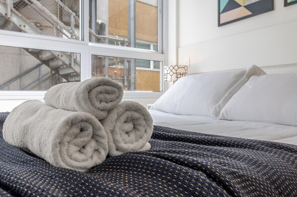 A neatly made bed with rolled towels and a patterned blanket, a window with a view of a building, and a geometric painting on the wall.