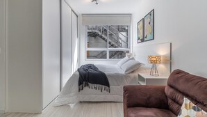 Ein modernes Schlafzimmer mit einem Bett, einem braunen Sessel, einem Nachttisch mit Lampe und einem Fenster mit Blick auf eine Treppe.