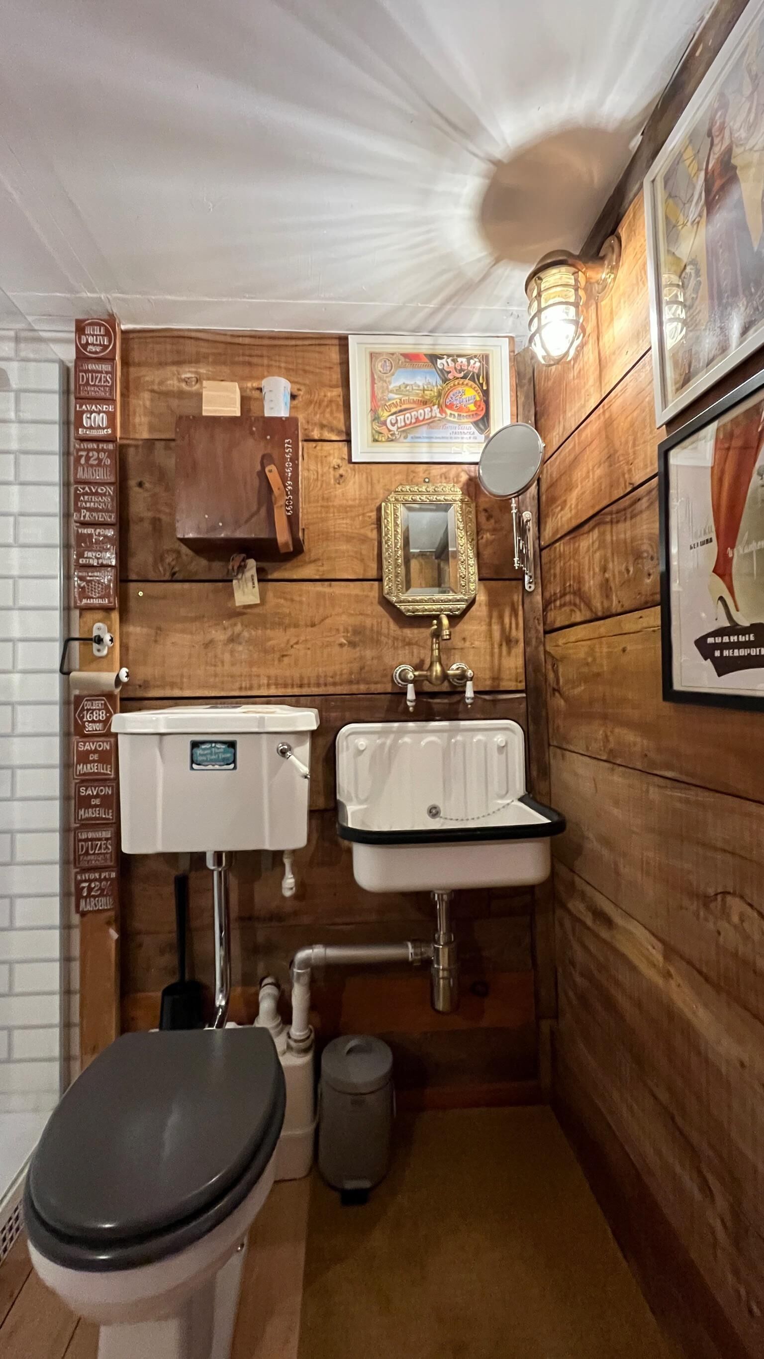 A compact bathroom with wooden walls, a white toilet, two sinks, a mirror, and a framed picture.