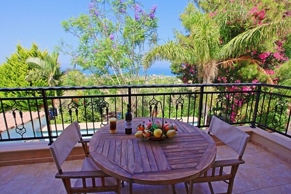A wooden table with chairs on a balcony with a view of the sea and lush greenery. - Marilena Sunset Villa Dio (Ayia Marina Chrysochous)
