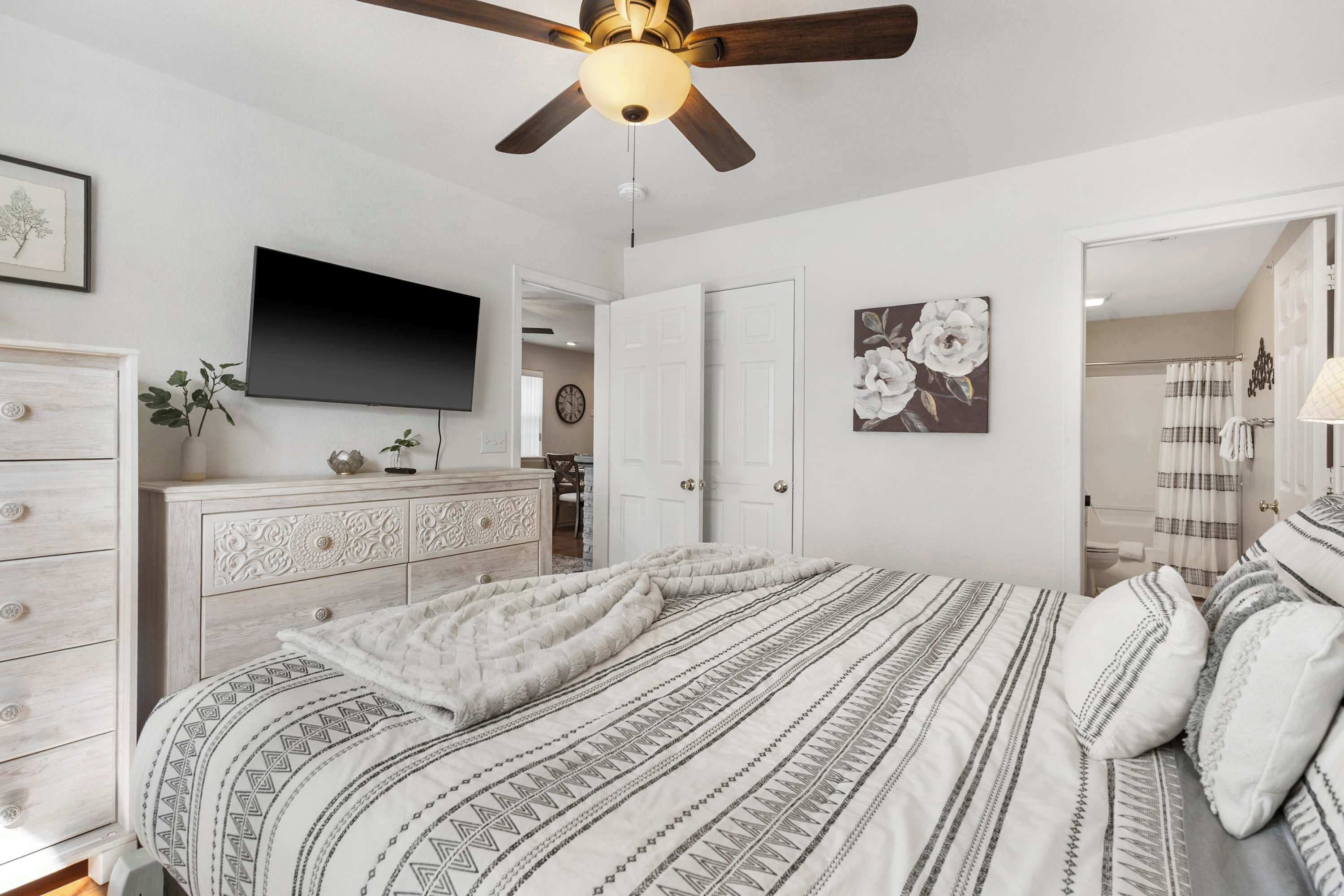 A bedroom with a bed, a ceiling fan, a TV, a dresser, and a door leading to another room.