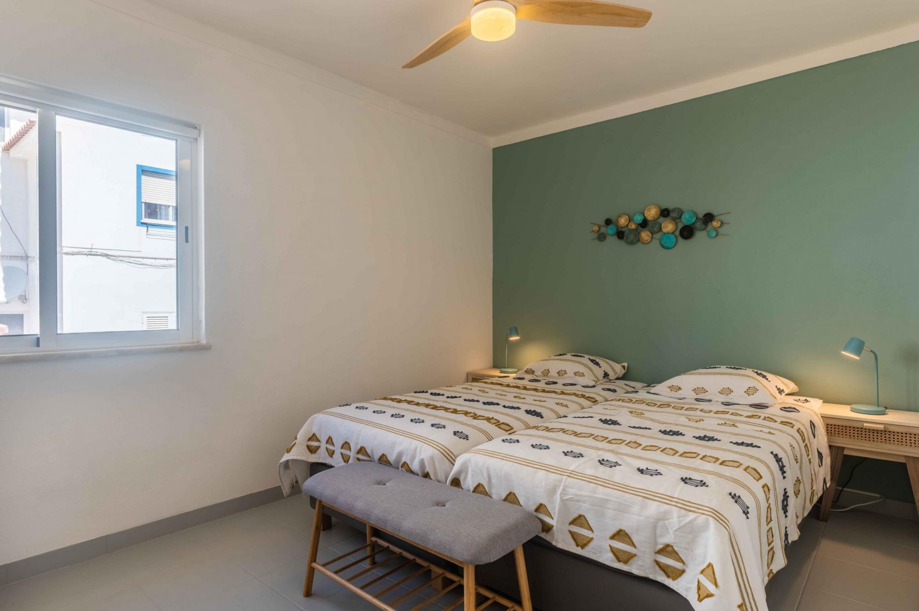 Un dormitorio con dos camas, un banco, un ventilador de techo, un adorno en la pared y una ventana.