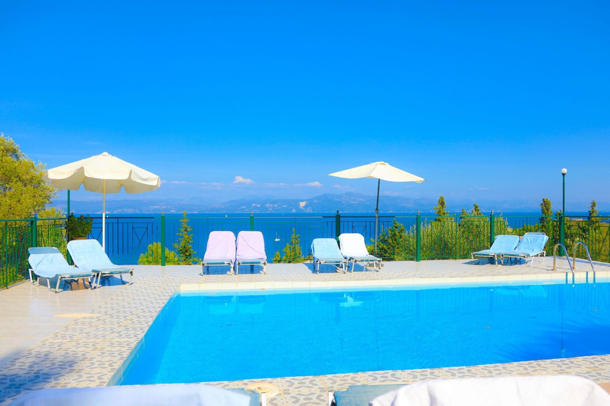 Uma área de piscina com espreguiçadeiras e guarda-sóis, com vista para um mar azul cristalino e montanhas ao longe.