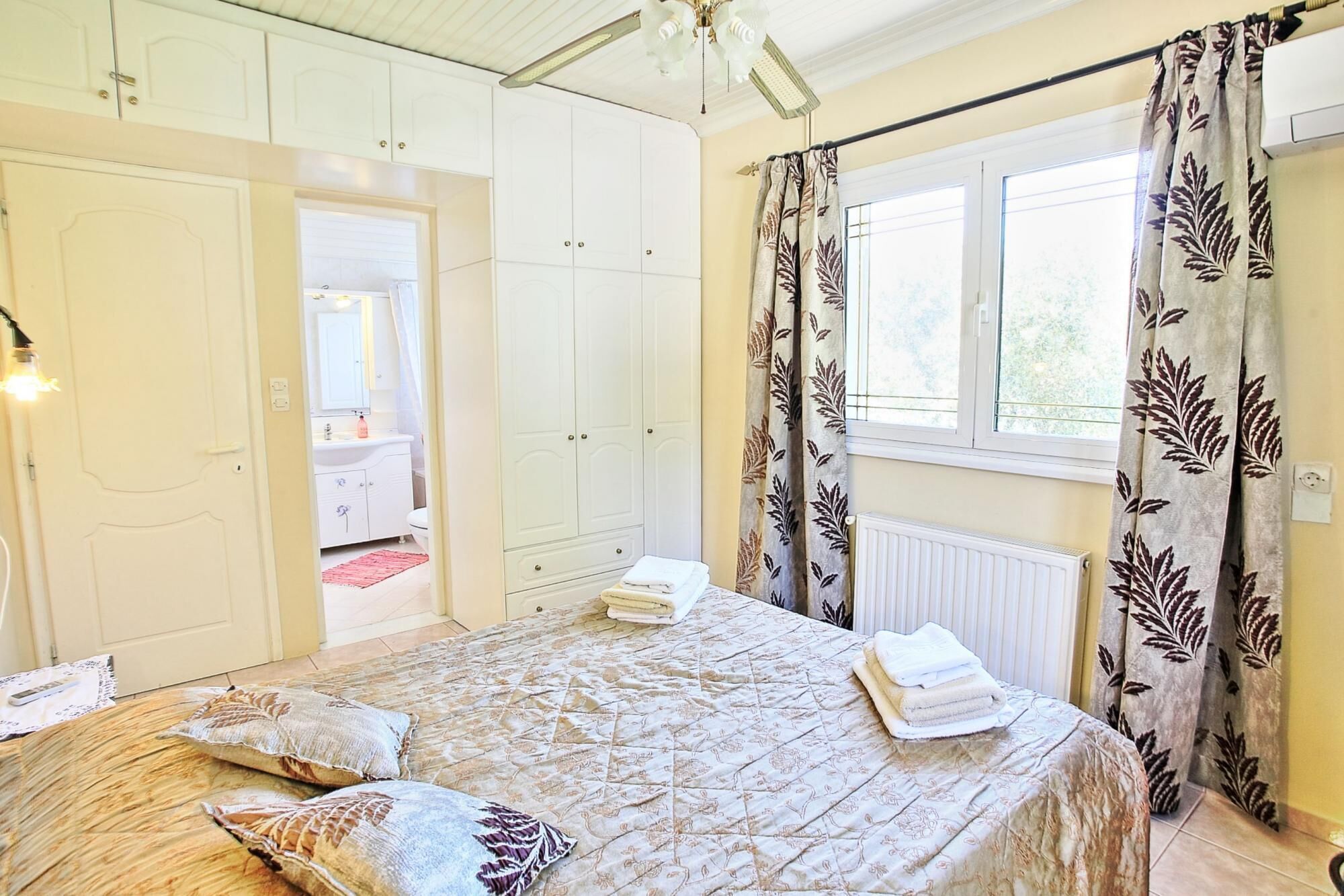 Ein Schlafzimmer mit Bett, Nachttisch, einem Fenster mit Vorhängen, einer Heizung und einem durch eine offene Tür einsehbaren Badezimmer.