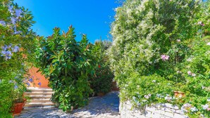 A stone-paved pathway leading through a lush garden with various flowering plants and a brick wall on one side. - Villa Ariadni in Nisaki (Corfu)