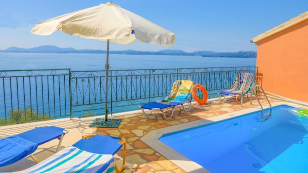 A pool area with lounge chairs, an umbrella, and a view of the sea. - Villa Ariadni in Nisaki (Corfu)
