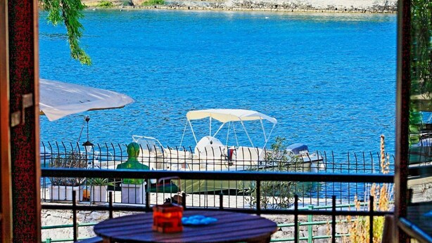 Eine Terrasse mit Tisch und Stühlen, mit Blick auf ein Gewässer, im Hintergrund Boote und Grünpflanzen.