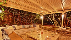 A covered outdoor seating area with a stone floor, a wooden table with candles, and a sofa with patterned cushions. - Prasini Luxury Cottage (Paxos)
