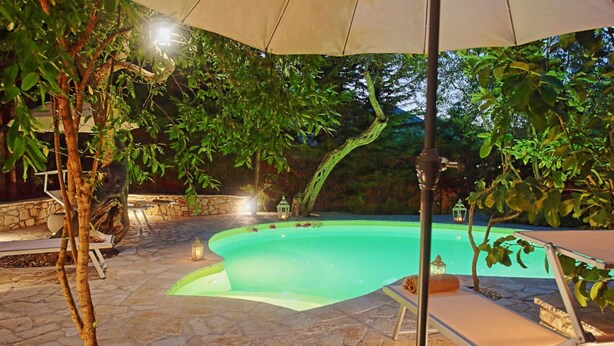 An outdoor swimming pool with stone flooring, surrounded by trees and a stone wall. - Prasini Luxury Cottage (Paxos)