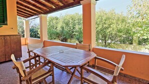 A covered outdoor patio with a wooden table and chairs, surrounded by a low wall and trees in the background.