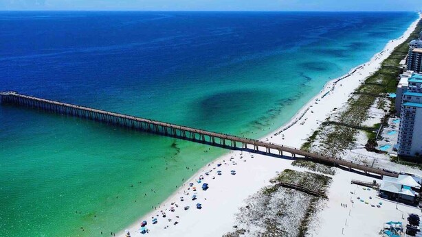 An aerial view of a beach with a long pier extending into the ocean, a sandy beach, and a building with a white facade and blue roof.