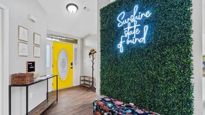 A modern hallway with a neon sign that reads "Sunshine state of mind," a yellow door with an oval window, and a wall covered in green foliage.
