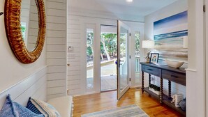A hallway with a wooden floor, a white door leading outside, a large mirror, and a painting of a beach scene. - Chic + Modern BHI Retreat, Both Clubs, 3 Golf Carts, 5 Porches w/Great Views (Bald Head Island)