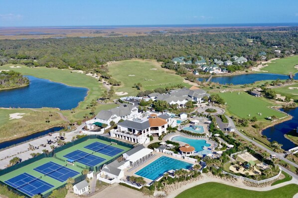 Vista aérea de un campo de golf con varios cuerpos de agua, una gran casa club, canchas de tenis y una piscina.