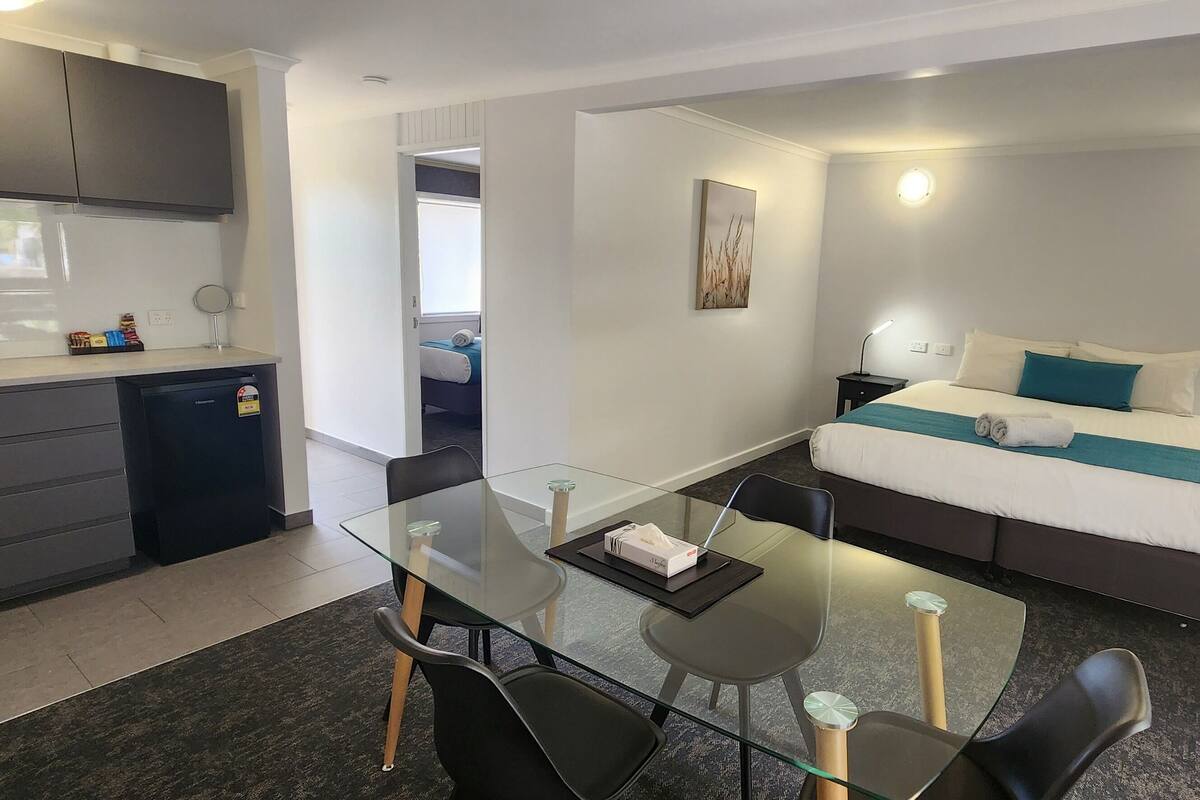 A modern hotel room with a glass dining table, a bed with a blue accent, a small kitchen area, and a bathroom visible through an open door.