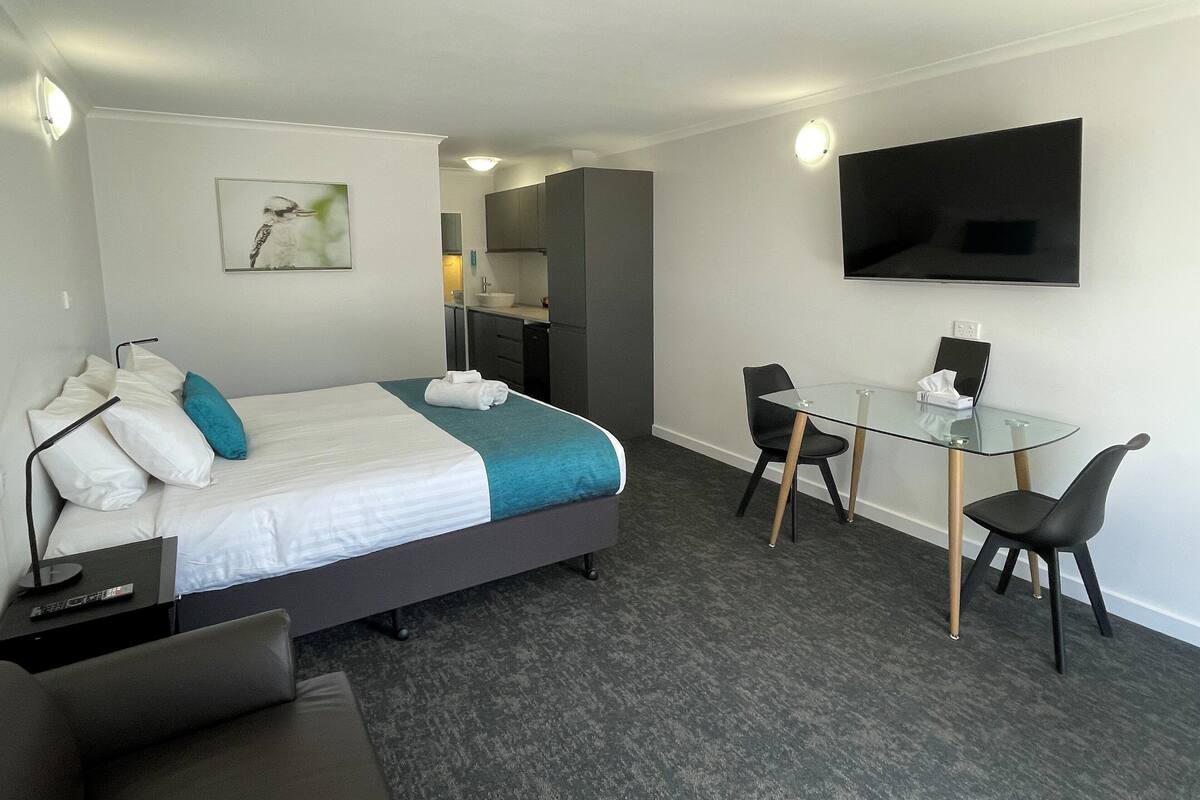 A modern hotel room with a bed, a desk, a chair, a TV, and a small kitchenette.