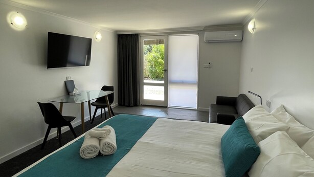 A hotel room with a bed, a desk, a chair, a TV, and a balcony with a view of greenery. - Barham Bridge Motor Inn (Barham)