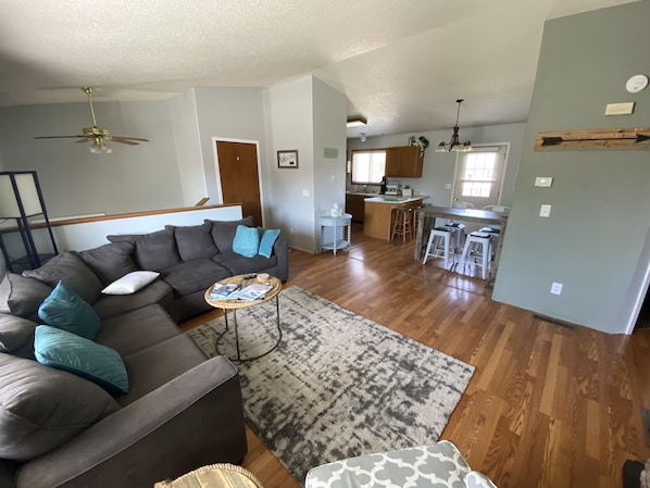A spacious living room with a sofa, a coffee table, and a patterned rug. A kitchen and dining area are visible in the background.