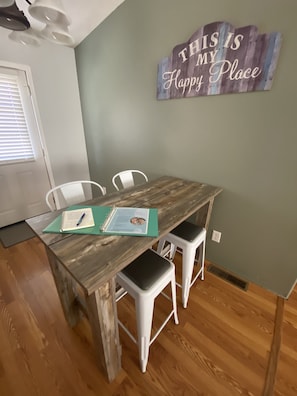 Un coin repas avec une table en bois, des chaises blanches et un panneau sur le mur où l’on peut lire : « ICI, C’EST MON PETIT BONHEUR ».
