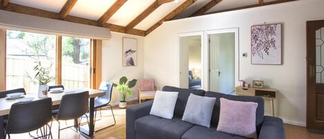 A modern living room with a dark grey sofa, wooden dining table, and chairs. There is a large window providing natural light and a view of the outdoors.