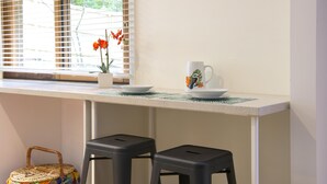 Una encimera de cocina con dos taburetes negros, una taza blanca con diseño de pájaro, una pequeña maceta con flores rojas y una cesta de mimbre con un paño de colores.