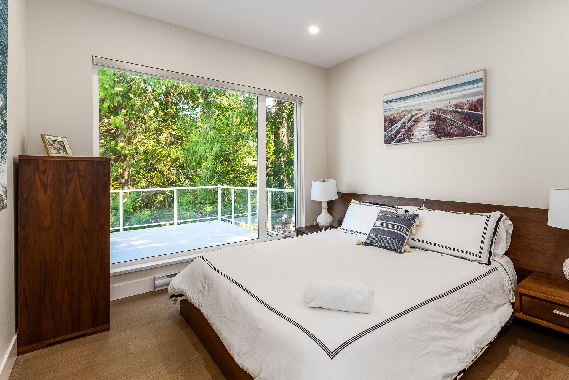Un dormitorio con una cama grande, una mesita de noche, un aparador de madera, un ventanal con vistas a la vegetación y un cuadro en la pared.