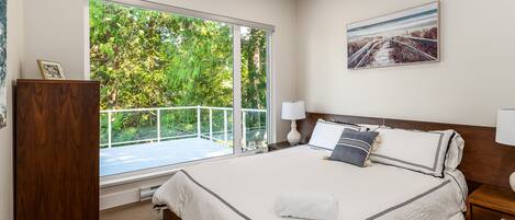 Un dormitorio con una cama grande, una mesita de noche, un aparador de madera, un ventanal con vistas a la vegetación y un cuadro en la pared.