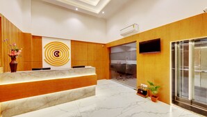 Un área de recepción moderna con un mostrador de mármol, paneles de madera, una gran obra de arte circular en la pared, un televisor de pantalla plana y un ascensor de vidrio.