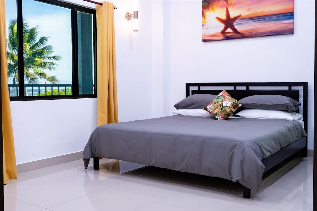 Une chambre à coucher avec un lit, une fenêtre avec des rideaux, un tableau accroché au mur et une vue sur des palmiers.