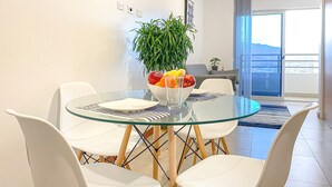 Un comedor con una mesa de vidrio, sillas blancas y un bol de fruta. Hay una planta en maceta y vistas a un balcón con un paisaje urbano.