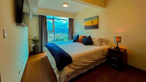 Un dormitorio con una cama grande, un televisor montado en la pared, una mesita de noche con lámpara y una ventana con vistas a la ciudad.