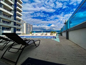 Zona de piscina en la azotea con sillones de descanso, vista al océano y un rascacielos al fondo.