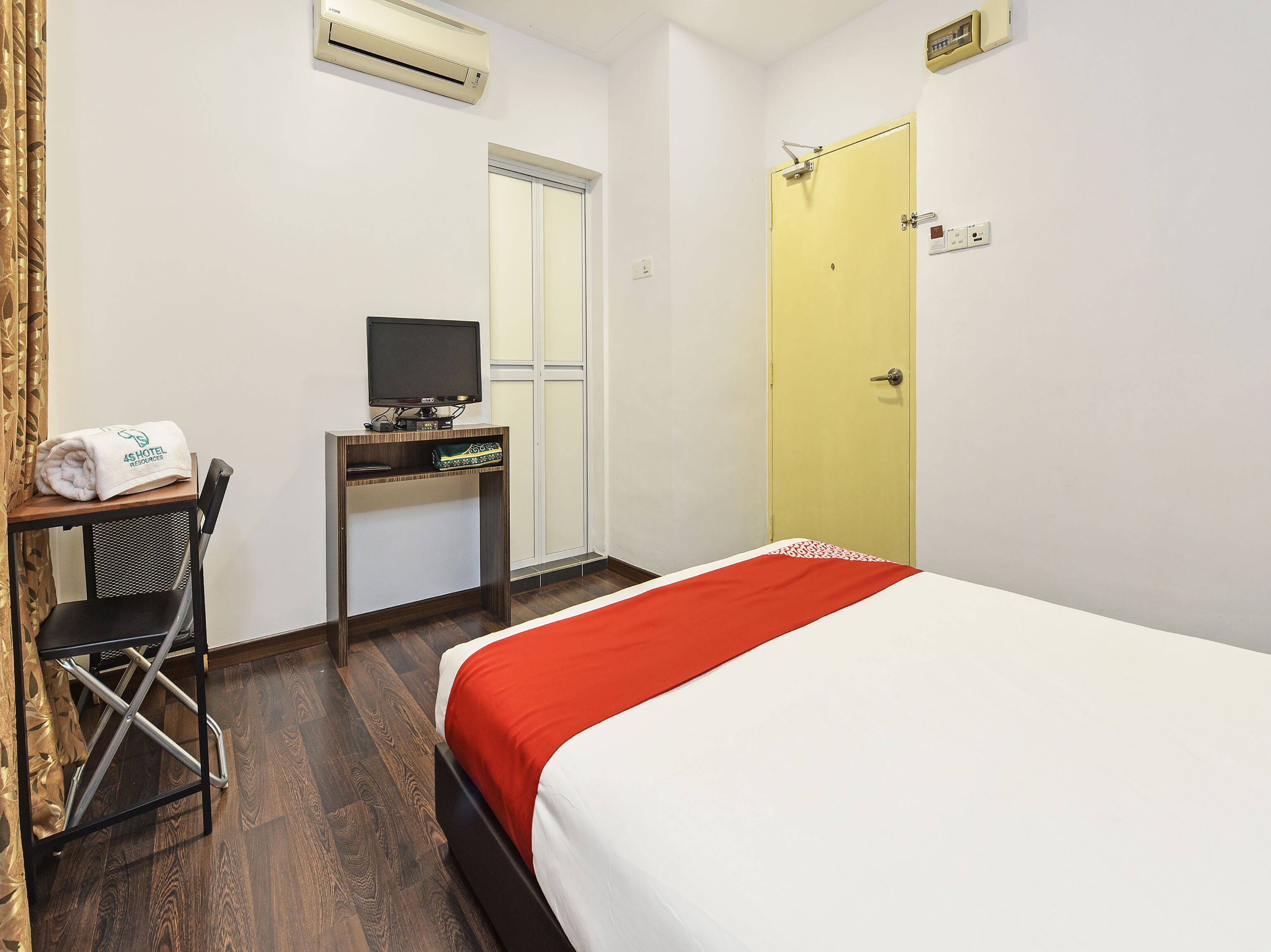 A hotel room with a bed, a desk with a television, a chair, an air conditioning unit, and a door.