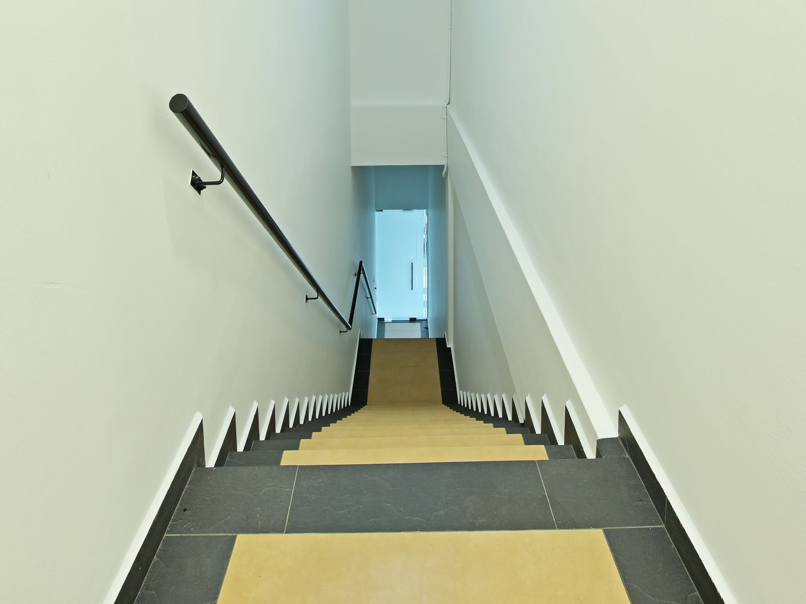 Una escalera con peldaños alternados en color negro y beige, balaustres blancos y barandilla negra.