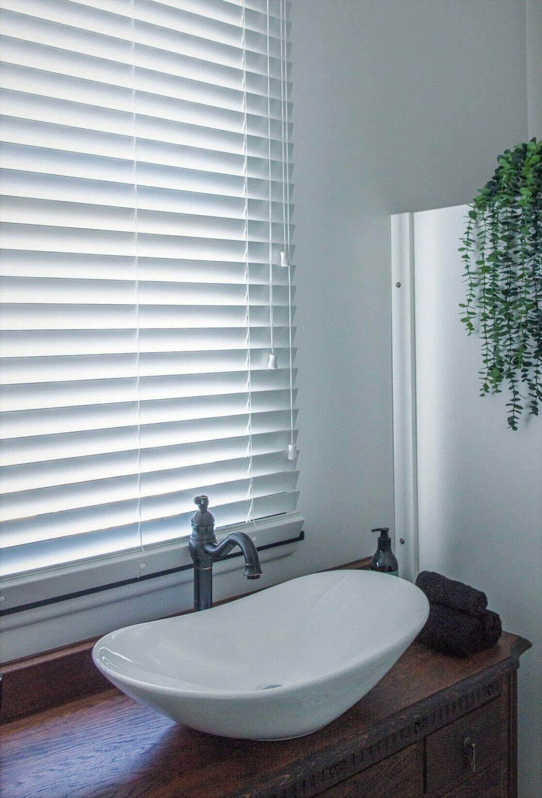 Un baño con lavamanos blanco, grifo, dispensador de jabón y ventana con persianas.
