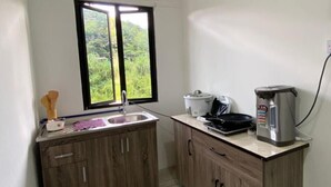A small kitchen with a sink, wooden cabinets, a window with a view of greenery, a kettle, and a coffee maker. - OYO Homes 90650 Na Homesuite (Ranau)