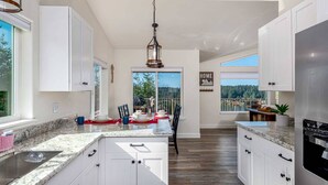 Una cocina amplia con armarios blancos, encimera de granito y una isla grande. Hay un comedor con sillas y una mesa dispuesta para dos personas. Un ventanal ofrece vistas al exterior.