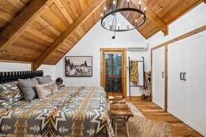 A bedroom with a sloped ceiling, a bed with patterned bedding, a chandelier, a door with a glass panel, and a wardrobe.