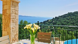 A balcony with a stone pillar, a table with a vase of flowers, a wooden chair, and a view of the sea and greenery. - Villa Regina in Kentroma (Corfu)
