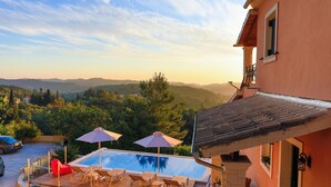A swimming pool with lounge chairs and umbrellas, surrounded by a wooden deck, with a view of hills and trees in the background. - Sunset House in Sidari (Corfu)
