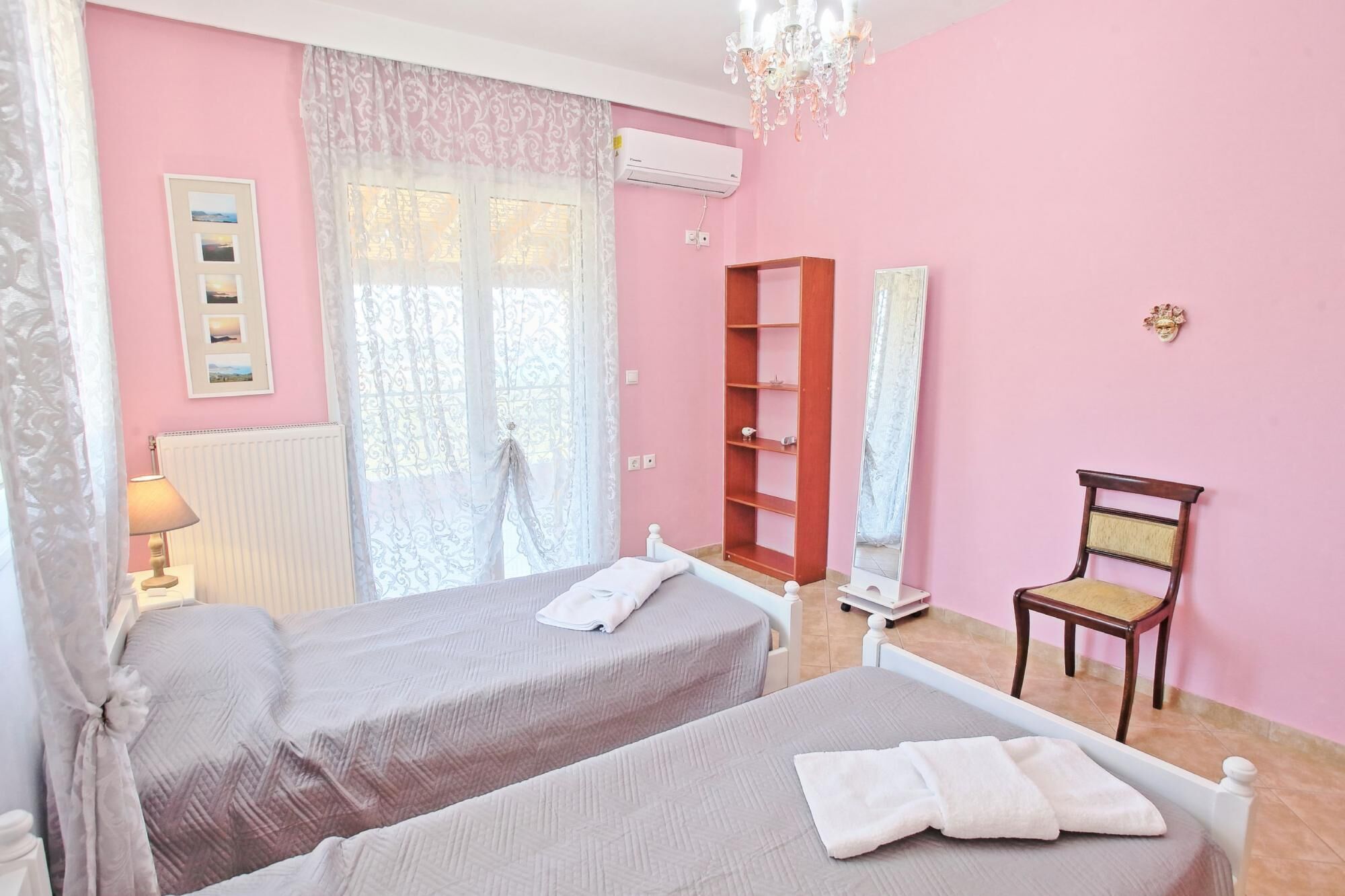 A hotel room with two beds, a wooden bookshelf, a chair, a mirror, and a chandelier.