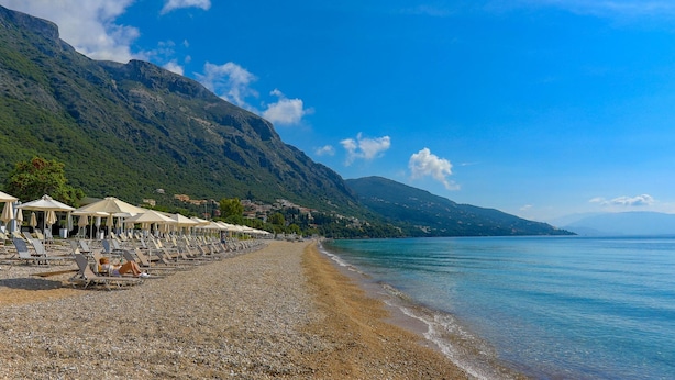 Une plage de galets avec des transats et des parasols, une mer d’un bleu limpide et des montagnes en arrière-plan.