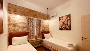 A room with two single beds, a stone wall, a window with curtains, a painting on the wall, and an air conditioning unit. - Mouragia House in Kerkira (Corfu)