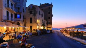 A street scene with outdoor seating at a restaurant, parked cars, and a building with balconies. - Mouragia House in Kerkira (Corfu)