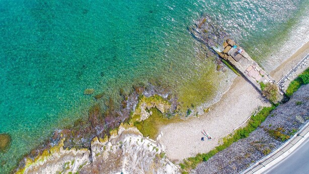 An aerial view of a coastal area with clear turquoise water, rocky shorelines, and a paved walkway along the beach. - Mouragia House in Kerkira (Corfu)